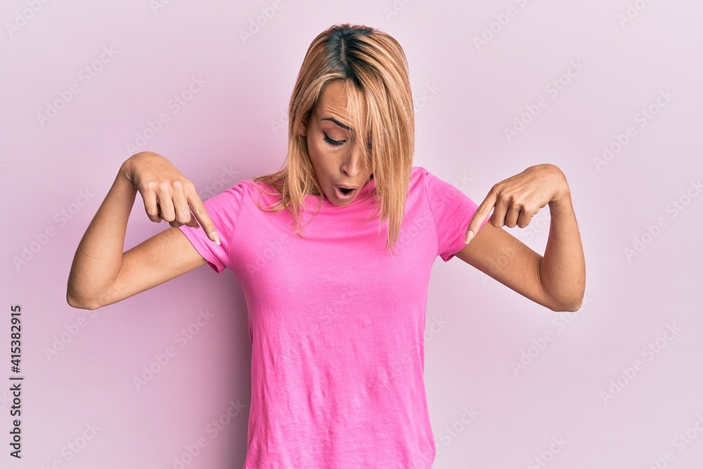 Beautiful blonde woman wearing casual pink tshirt pointing down with fingers showing advertisement, surprised face and open mouth Beautiful blonde woman wearing casual pink tshirt pointing down with fingers showing advertisement, surprised face and open mouth