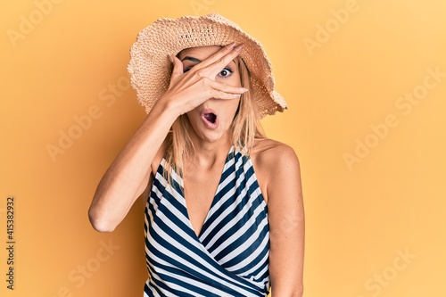 Beautiful blonde woman wearing summer hat peeking in shock covering face and eyes with hand, looking through fingers afraid