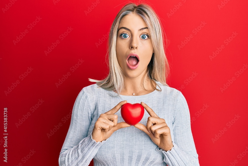 Beautiful blonde woman holding heart afraid and shocked with surprise and amazed expression, fear and excited face. Beautiful blonde woman holding heart afraid and shocked with surprise and amazed expression, fear and excited face.