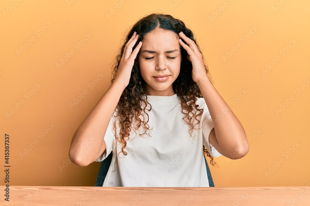 Teenager hispanic girl wearing casual clothes sitting on the table with hand on head for pain in head because stress. suffering migraine.