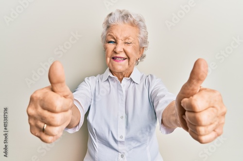 Senior grey-haired woman doing thumbs up positive gesture winking looking at the camera with sexy expression, cheerful and happy face.