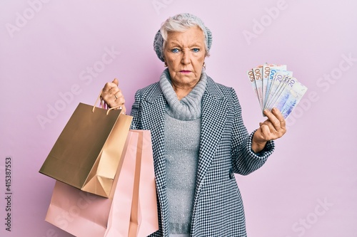 Senior grey-haired woman holding shopping bags and swedish krona banknotes depressed and worry for distress, crying angry and afraid. sad expression.