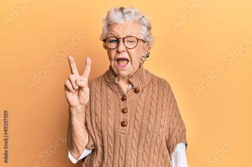 Foto Senior grey-haired woman wearing casual clothes and glasses smiling with happy face winking at the camera doing victory sign