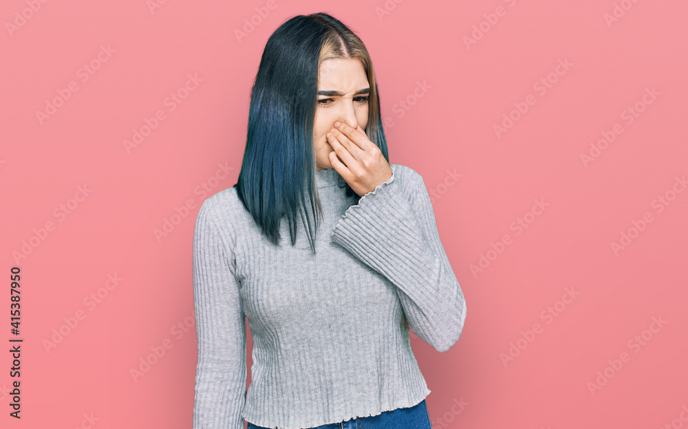 Young modern girl wearing casual sweater smelling something stinky and disgusting, intolerable smell, holding breath with fingers on nose. bad smell