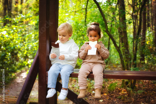 A little blonde boy and an African-American girl in a park on a bench eating a bar of white and dark chocolate. © Elena 