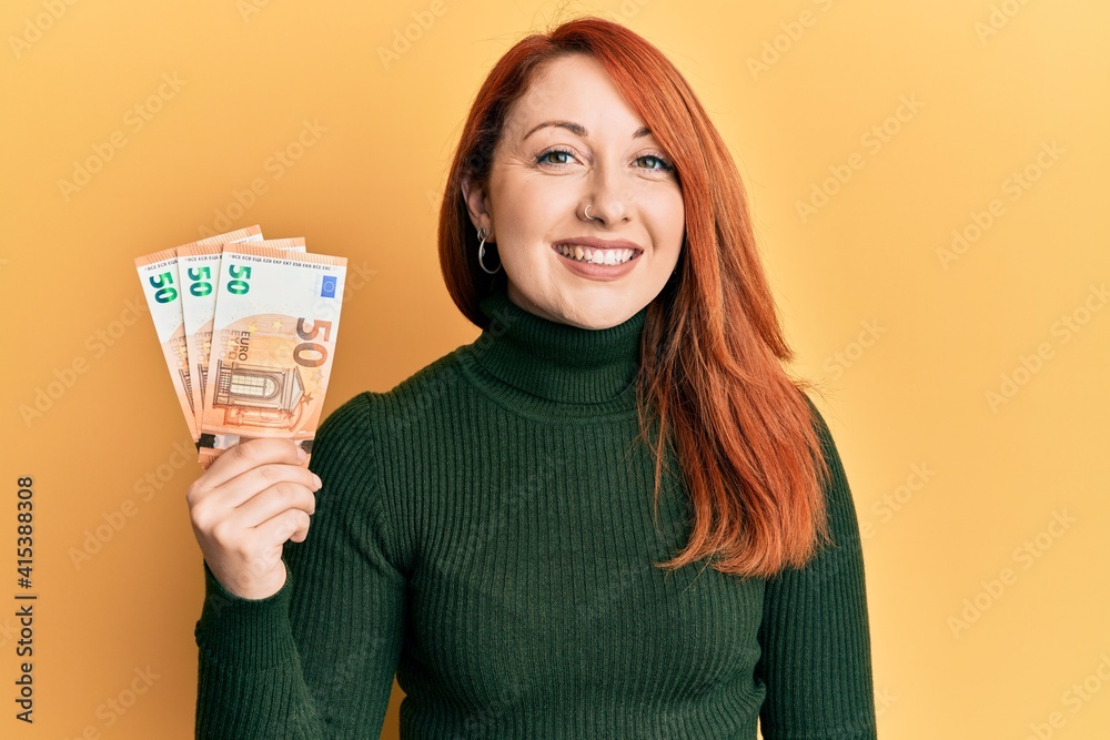 Beautiful redhead woman holding bunch of 50 euro banknotes looking positive and happy standing and smiling with a confident smile showing teeth Beautiful redhead woman holding bunch of 50 euro banknotes looking positive and happy standing and smiling with a confident smile showing teeth