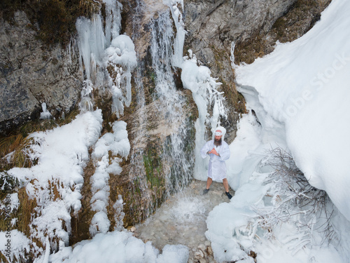 European man in traditional Japanese shugendo outfit meditating next to waterfall surrounded by icicles and snow in winter in austria photo