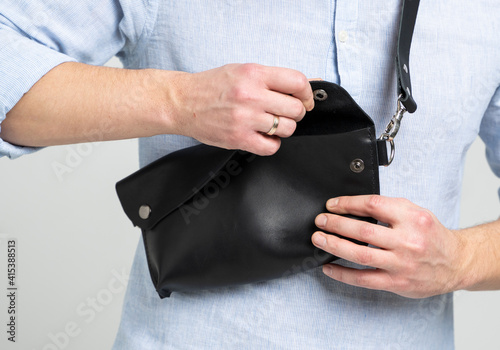 Man in a blue shirt with a leather black handmade bag over his shoulder. Dark designer banana bag. Man in a studio. Comfortable small bag for walking.