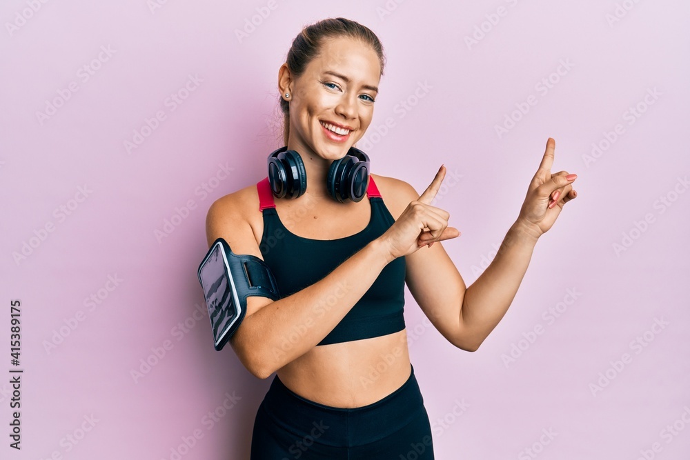 Beautiful young blonde woman wearing gym clothes and using headphones smiling and looking at the camera pointing with two hands and fingers to the side. Beautiful young blonde woman wearing gym clothes and using headphones smiling and looking at the camera pointing with two hands and fingers to the side.