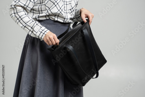 Small black leather bag in a woman's hand on a white background. Shoulder handbag. Woman in a white plaid shirt and black jeans and with a black handbag. Style, retro, fashion, vintage and elegance.
