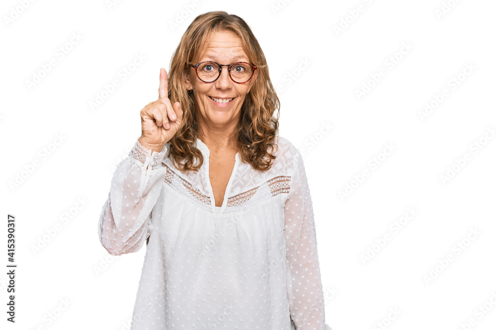 Middle age blonde woman wearing casual white shirt and glasses showing and pointing up with finger number one while smiling confident and happy.