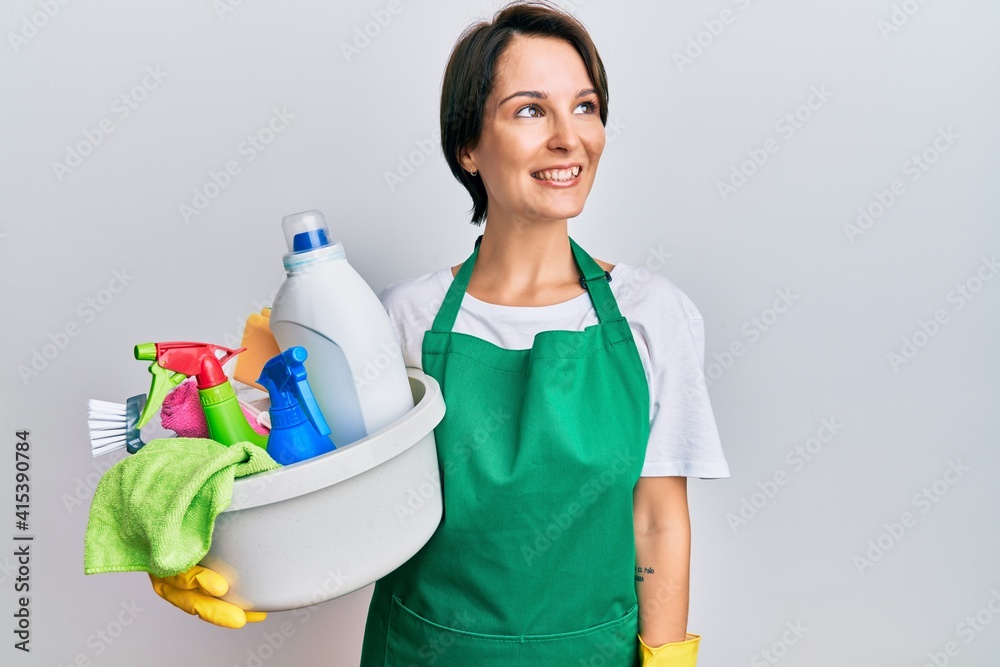 Young brunette woman with short hair wearing apron holding cleaning products smiling looking to the side and staring away thinking. Young brunette woman with short hair wearing apron holding cleaning products smiling looking to the side and staring away thinking.