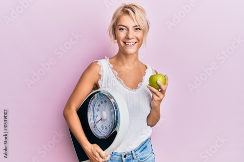 Young blonde girl holding weight machine and green apple smiling with a happy and cool smile on face. showing teeth.