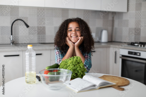 Young beautiful mixed race curly girl sitting in the kitchen at the table  putting her head on her hands and smiling. Healthy beverage  natural hydration body balance concept