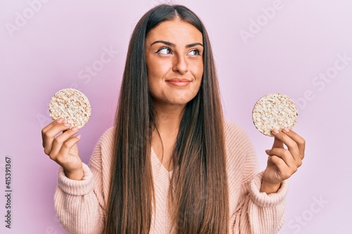 Beautiful hispanic woman eating healthy rice crackers smiling looking to the side and staring away thinking.