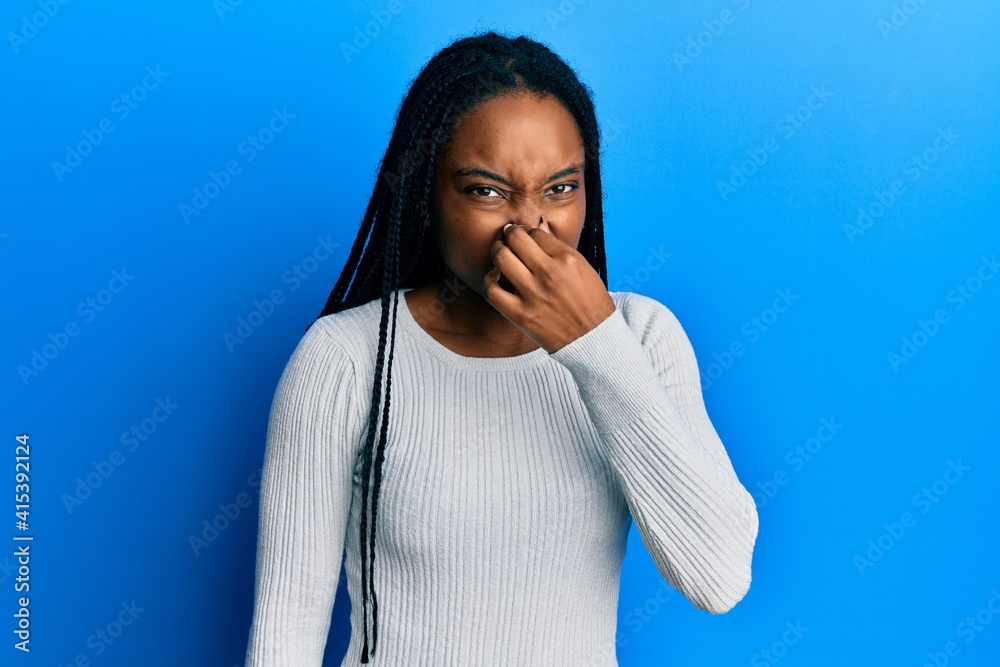 African american woman with braided hair wearing casual white sweater smelling something stinky and disgusting, intolerable smell, holding breath with fingers on nose. bad smell