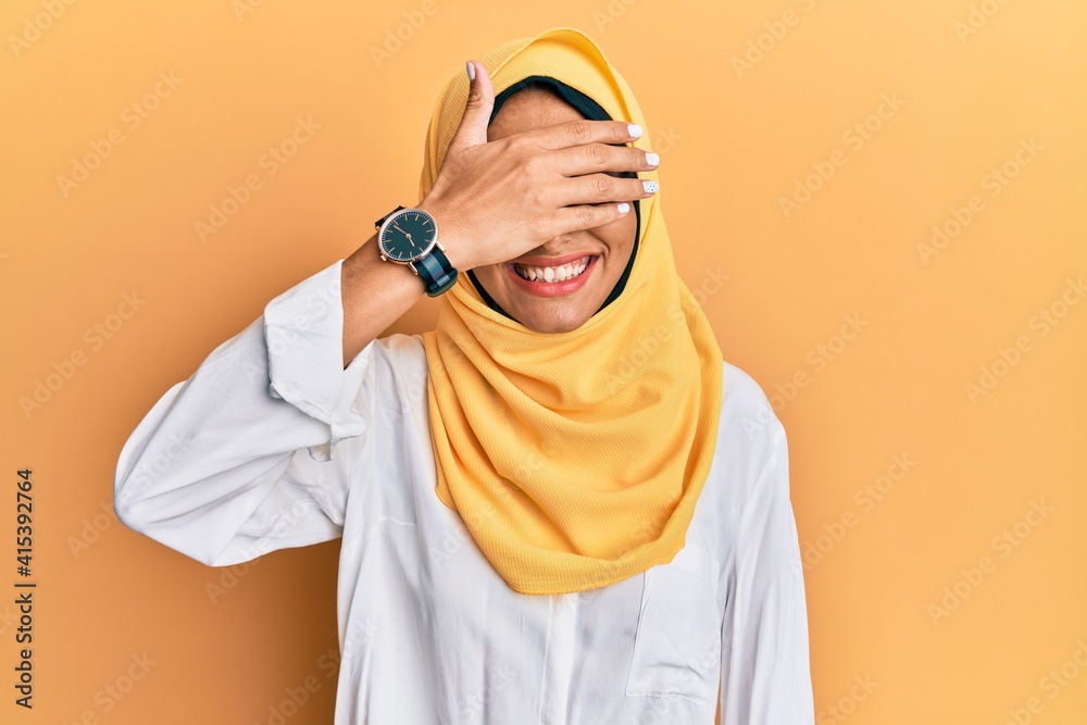 Young brunette arab woman wearing traditional islamic hijab scarf smiling and laughing with hand on face covering eyes for surprise. blind concept. Young brunette arab woman wearing traditional islamic hijab scarf smiling and laughing with hand on face covering eyes for surprise. blind concept.