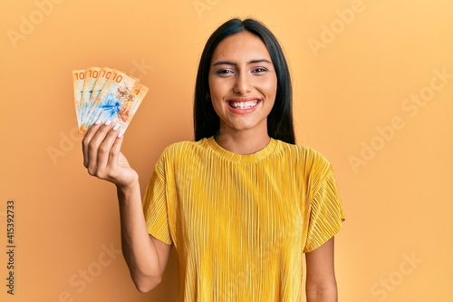 Young brunette woman holding 10 swiss franc banknotes looking positive and happy standing and smiling with a confident smile showing teeth