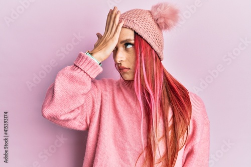 Young caucasian woman wearing wool sweater and winter hat surprised with hand on head for mistake, remember error. forgot, bad memory concept.