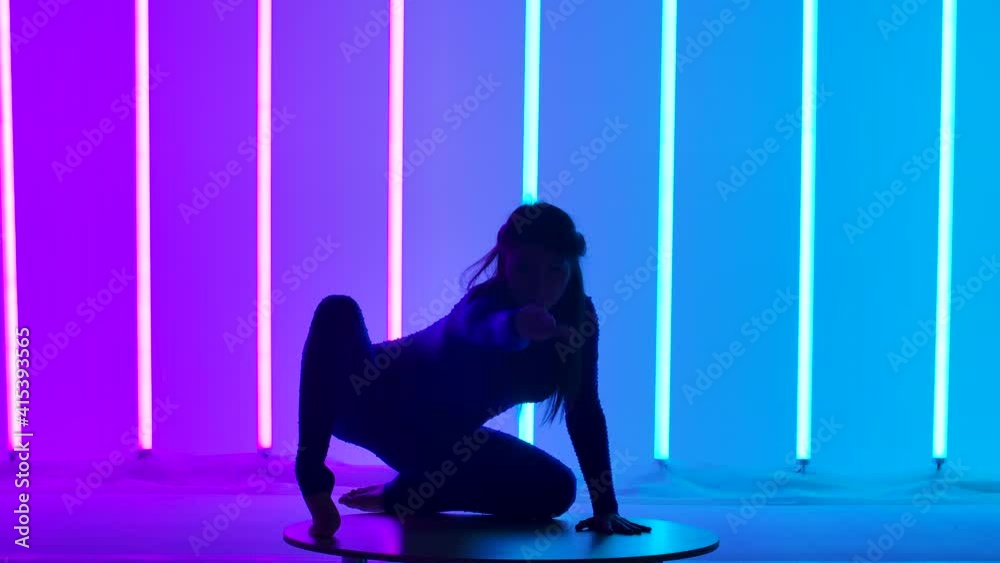 An attractive woman performs a handstand, moves her legs and splits them into a split. Gymnast in the studio against the background of pink and blue lights. Silhouette. Slow motion. Close up.