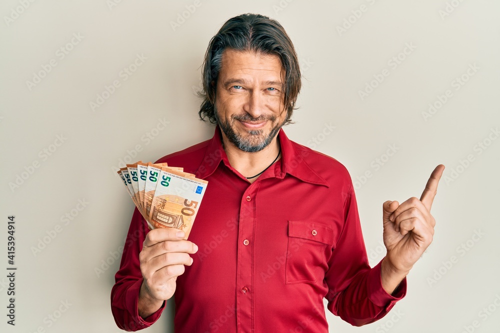 Middle age handsome man holding bunch of 50 euro banknotes smiling happy pointing with hand and finger to the side Middle age handsome man holding bunch of 50 euro banknotes smiling happy pointing with hand and finger to the side