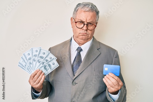 Senior grey-haired man wearing business suit holding credit car and dollars depressed and worry for distress, crying angry and afraid. sad expression.