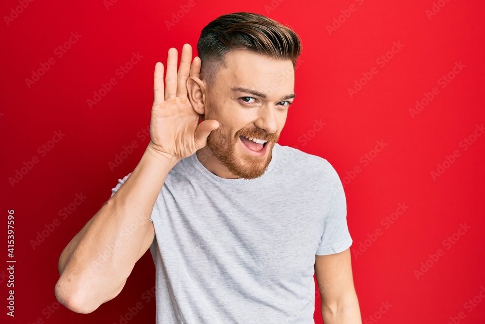 Young redhead man wearing casual grey t shirt smiling with hand over ear listening and hearing to rumor or gossip. deafness concept. Young redhead man wearing casual grey t shirt smiling with hand over ear listening and hearing to rumor or gossip. deafness concept.