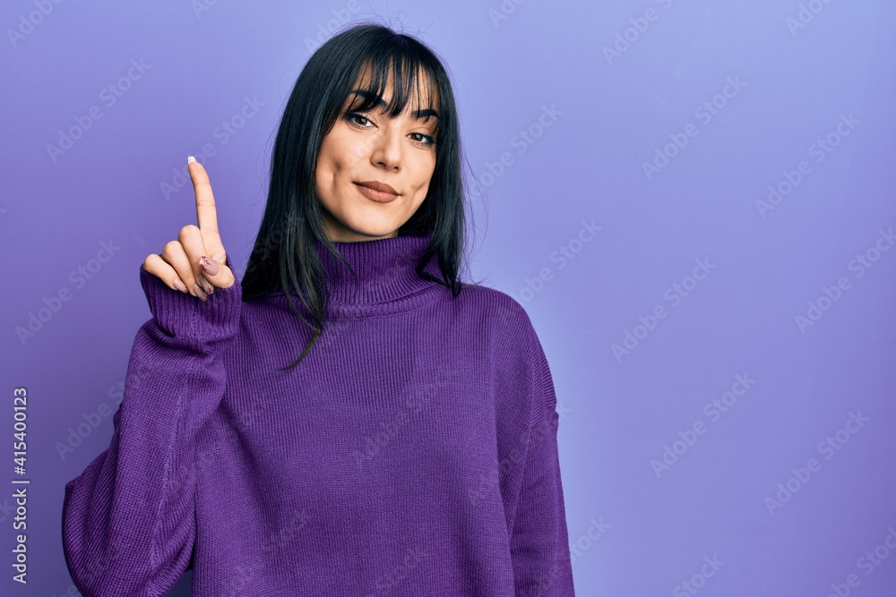 Young brunette woman with bangs wearing turtleneck sweater showing and pointing up with finger number one while smiling confident and happy. Young brunette woman with bangs wearing turtleneck sweater showing and pointing up with finger number one while smiling confident and happy.