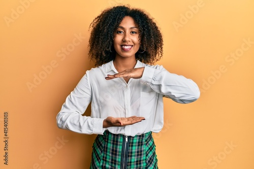 Beautiful african american woman with afro hair wearing scholar skirt gesturing with hands showing big and large size sign, measure symbol. smiling looking at the camera. measuring concept.