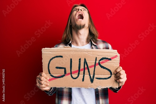 Handsome caucasian man with long hair holding no guns warning banner angry and mad screaming frustrated and furious, shouting with anger looking up.