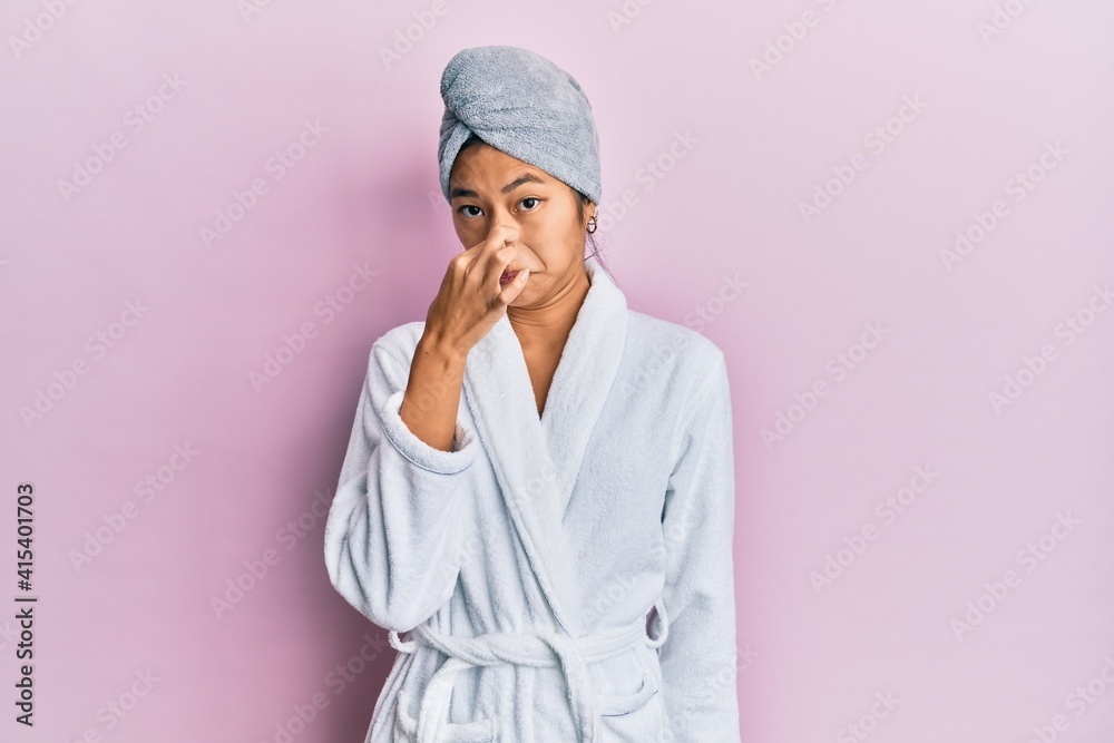 Young chinese woman wearing shower towel cap and bathrobe smelling something stinky and disgusting, intolerable smell, holding breath with fingers on nose. bad smell Young chinese woman wearing shower towel cap and bathrobe smelling something stinky and disgusting, intolerable smell, holding breath with fingers on nose. bad smell