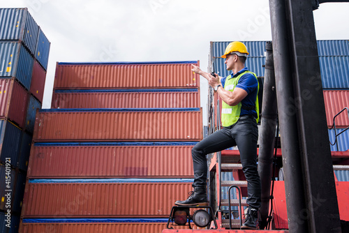 Confident Caucasian man engineer using radio communication or walkies talkie and wearing yellow safety helmet and check for control loading containers box from Cargo freight ship for import and export