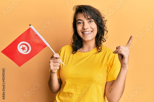 Young hispanic woman holding tunisia flag smiling happy pointing with hand and finger to the side