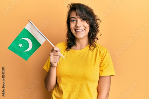 Young hispanic woman holding pakistan flag looking positive and happy standing and smiling with a confident smile showing teeth