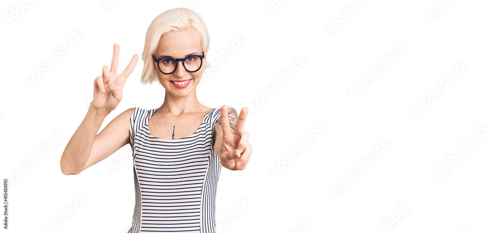 Young blonde woman with tattoo wearing casual clothes and glasses smiling looking to the camera showing fingers doing victory sign. number two.