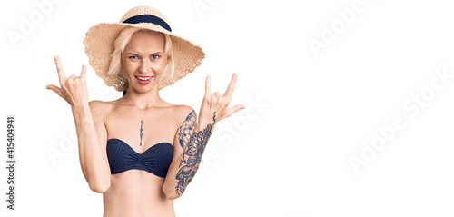 Young blonde woman with tattoo wearing bikini and summer hat shouting with crazy expression doing rock symbol with hands up. music star. heavy concept.