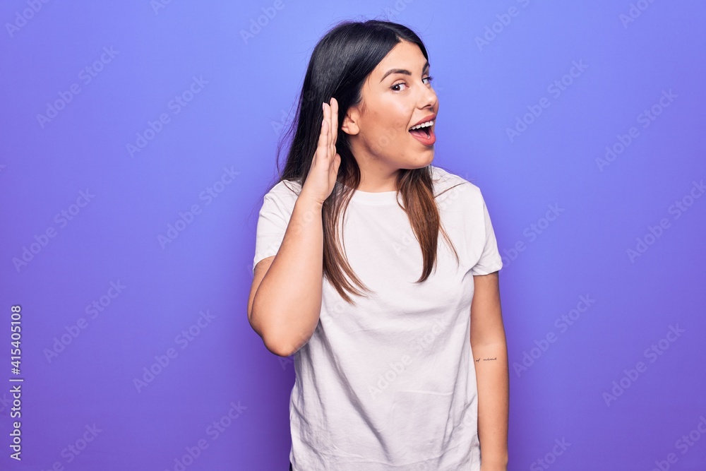 Young beautiful brunette woman wearing casual t-shirt standing over purple background smiling with hand over ear listening and hearing to rumor or gossip. Deafness concept. Young beautiful brunette woman wearing casual t-shirt standing over purple background smiling with hand over ear listening and hearing to rumor or gossip. Deafness concept.