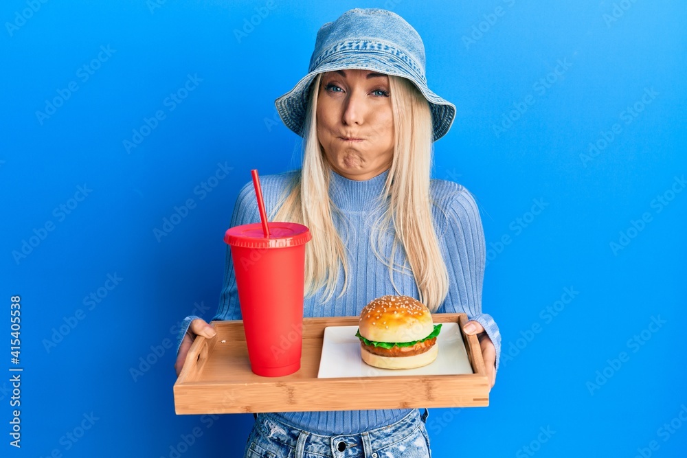 Young blonde woman eating a tasty classic burger and soda puffing cheeks with funny face. mouth inflated with air, catching air.
