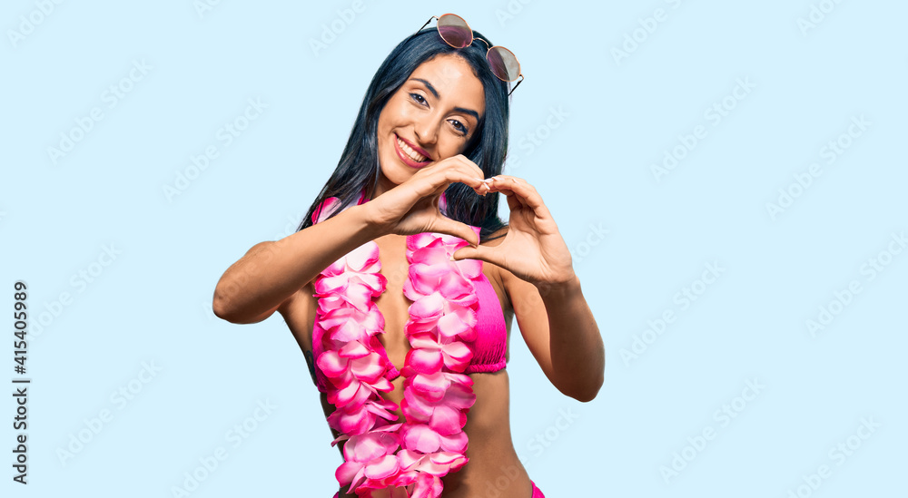 Beautiful hispanic woman wearing bikini and hawaiian lei smiling in love doing heart symbol shape with hands. romantic concept. Beautiful hispanic woman wearing bikini and hawaiian lei smiling in love doing heart symbol shape with hands. romantic concept.