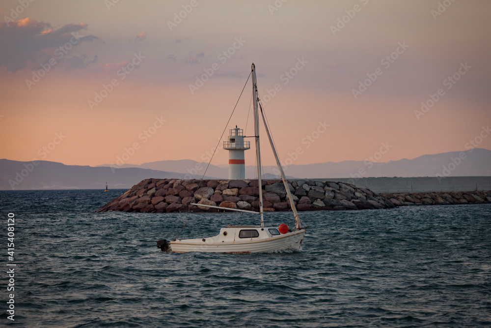 Sailboat is sailing towards the bay and the lighthouse is visible among the mast at dusk with reddish sky. Wavy sea, lighthouse and small sailboat sailing during sunset. Holiday concept.