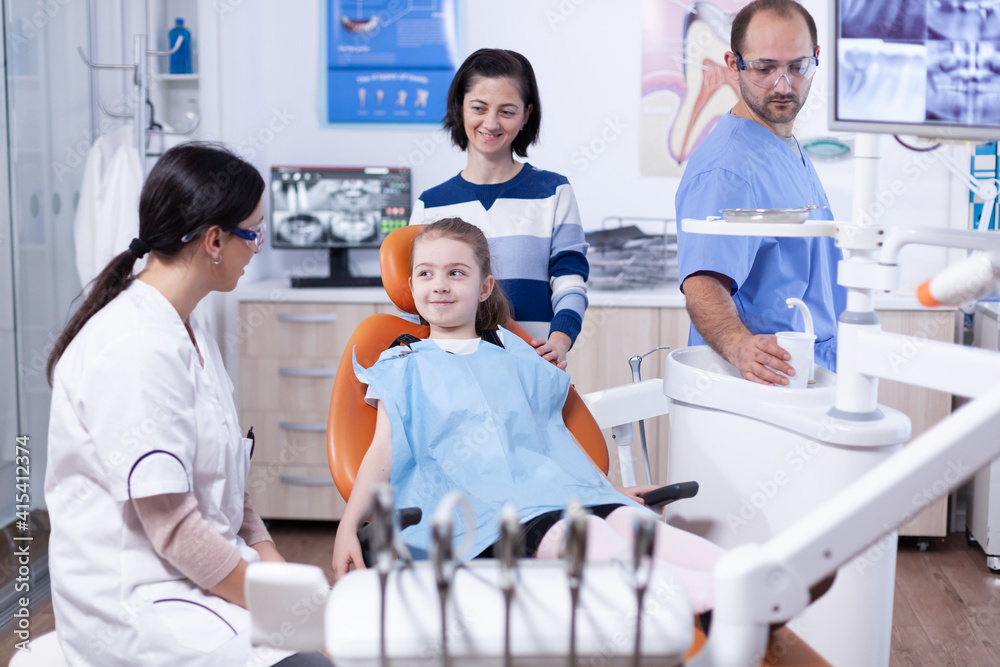 Obraz premium Happy little girl at pediatric dentist wearing dental bib during stomatology examination. Child with her mother during teeth check up with stomatolog sitting on chair.
