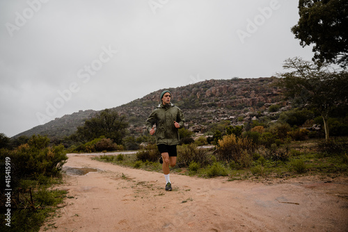 Handsome young male sprinter training in cloudy weather running on gravel mountain path with green bushes and hills