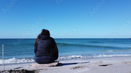 Beautiful woman sitting on the shore of a blue sea from behind on a windy day. Relaxing Concept. Young woman sits on beach and looks at beautiful landscape with sea