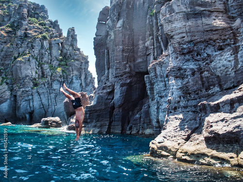 Unrecognizable shirtless man plunging into blue sea water near rocky cliff on summer day in nature