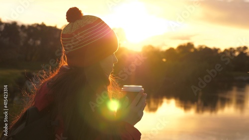 Woman Traveler holds mug with hot coffee in her hands and looks at sunset byriver. A tourist drinks tea from mug in sun. Free traveler girl admiring scenery. Hiking. Hiker drinking hot cocoa