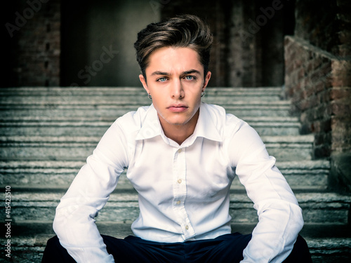 One handsome elegant young man in urban setting in European city, sitting on old stairs, looking at camera