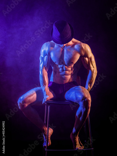 Wallpaper Mural Shirtless muscular bodybuilder performer sitting on stool wearing hat in studio