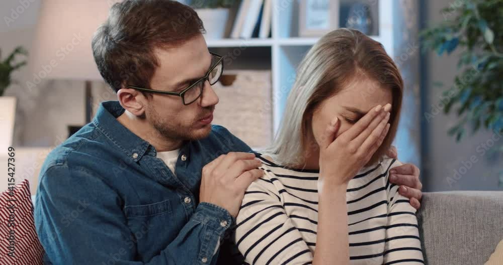 Young couple sitting at home on couch talking. Husband holds his wife by shoulders, who is crying after receiving bad news. Young couple sitting at home on couch talking. Husband holds his wife by shoulders, who is crying after receiving bad news.