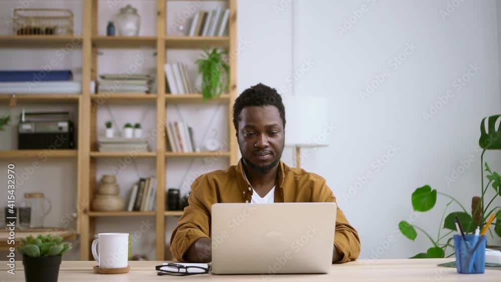 Businessman working with computer remotely and sitting at table in home office spbas. Young American man opens laptop, looks at screen and sits at desk in light room. Front view of millennial