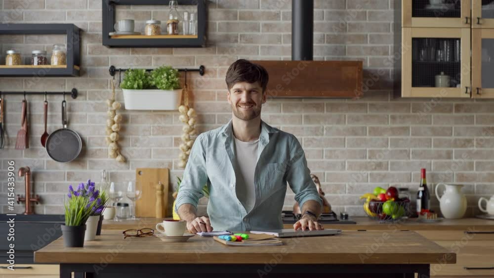 Lockdown medium shot portrait of young man finishing remote work, closing laptop computer and smiling at camera standing at table in domestic kitchen